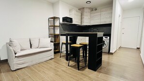 Un espacio habitable compacto con sofá, una pequeña barra con taburetes, lavadora y cocina integrada con armarios.