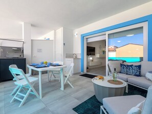 Ein modernes Wohnzimmer mit einer Couch, einem Esstisch mit Stühlen, einer Küchenzeile inklusive Mikrowelle und Meerblick durch eine Schiebetür.