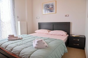 Una cama bien tendida con una colcha verde y rosa, una pila de toallas dobladas y una mesita de noche con un libro.