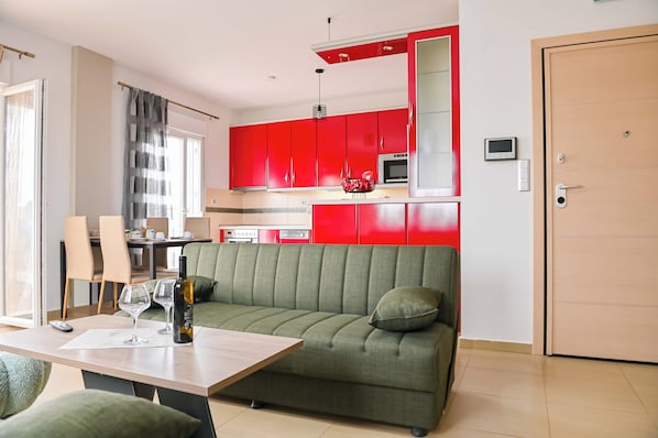 A modern living room with a green sofa, a dining table with chairs, and a kitchen area with red cabinets.