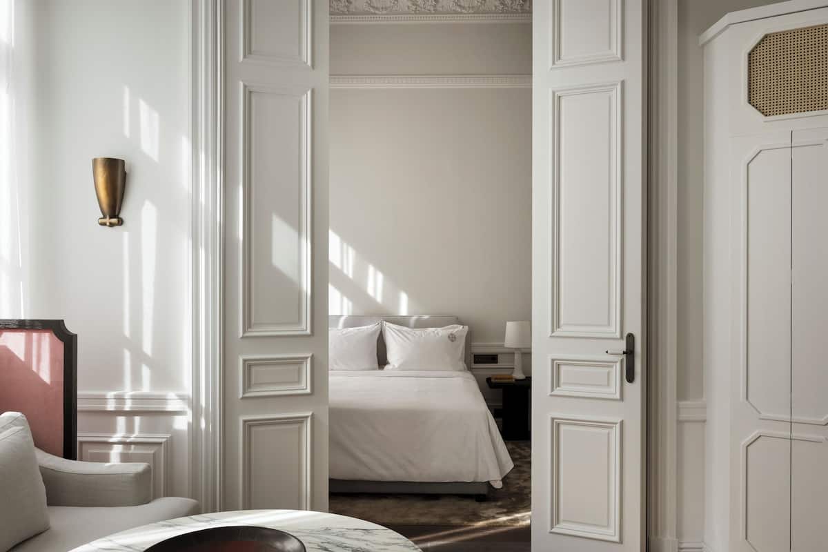 Quarto de hotel com uma cama, mesa de cabeceira, luminária e porta com detalhe decorativo.