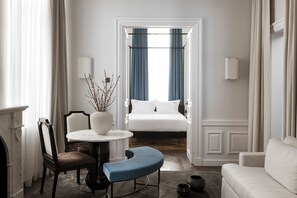 Quarto de hotel com uma cama, uma mesa redonda com cadeiras, um banco azul, uma lareira e vista através de uma porta aberta.