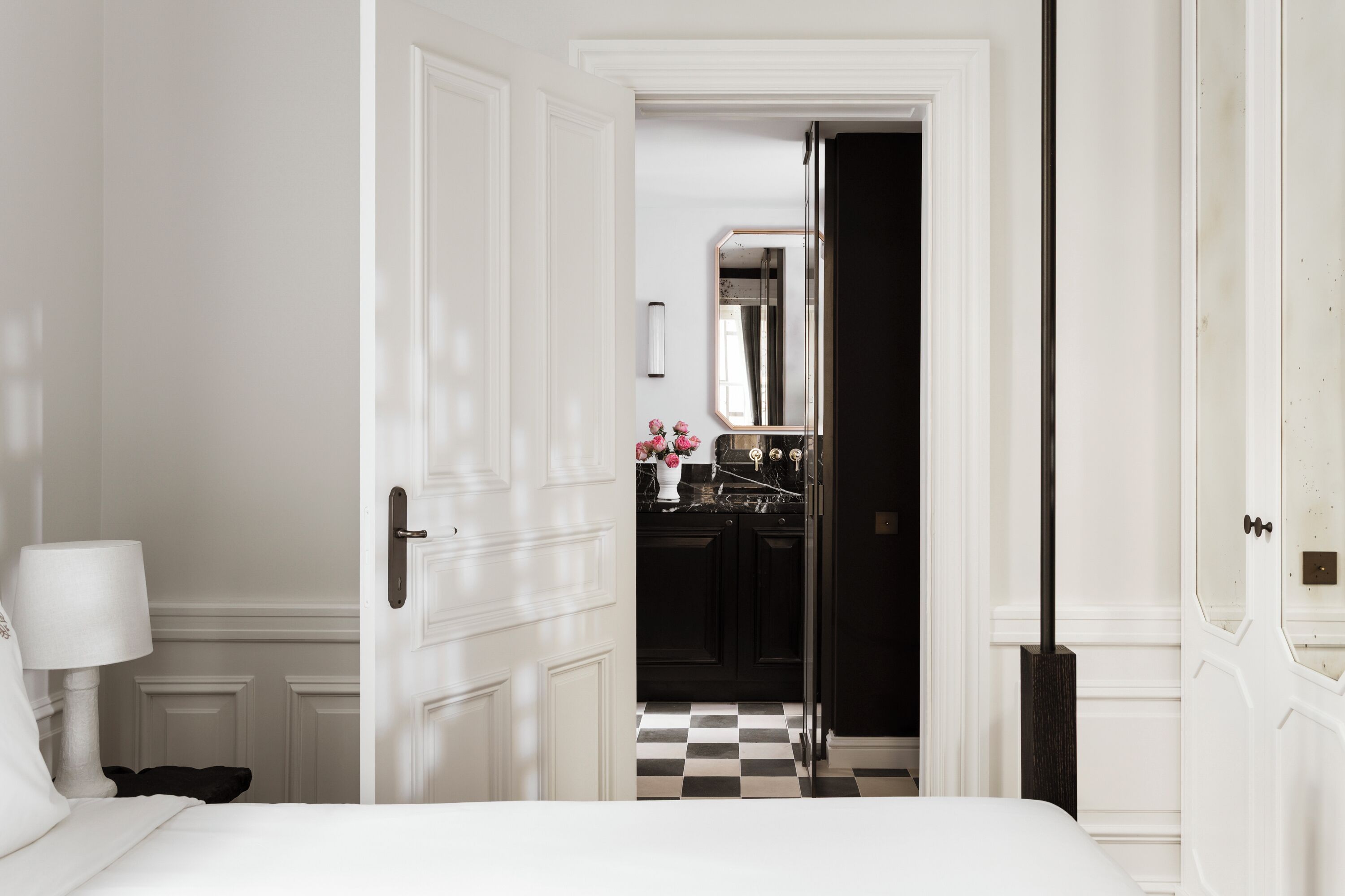 A hotel room with a checkered floor, a bed, a nightstand, a lamp, and a door leading to a bathroom.
