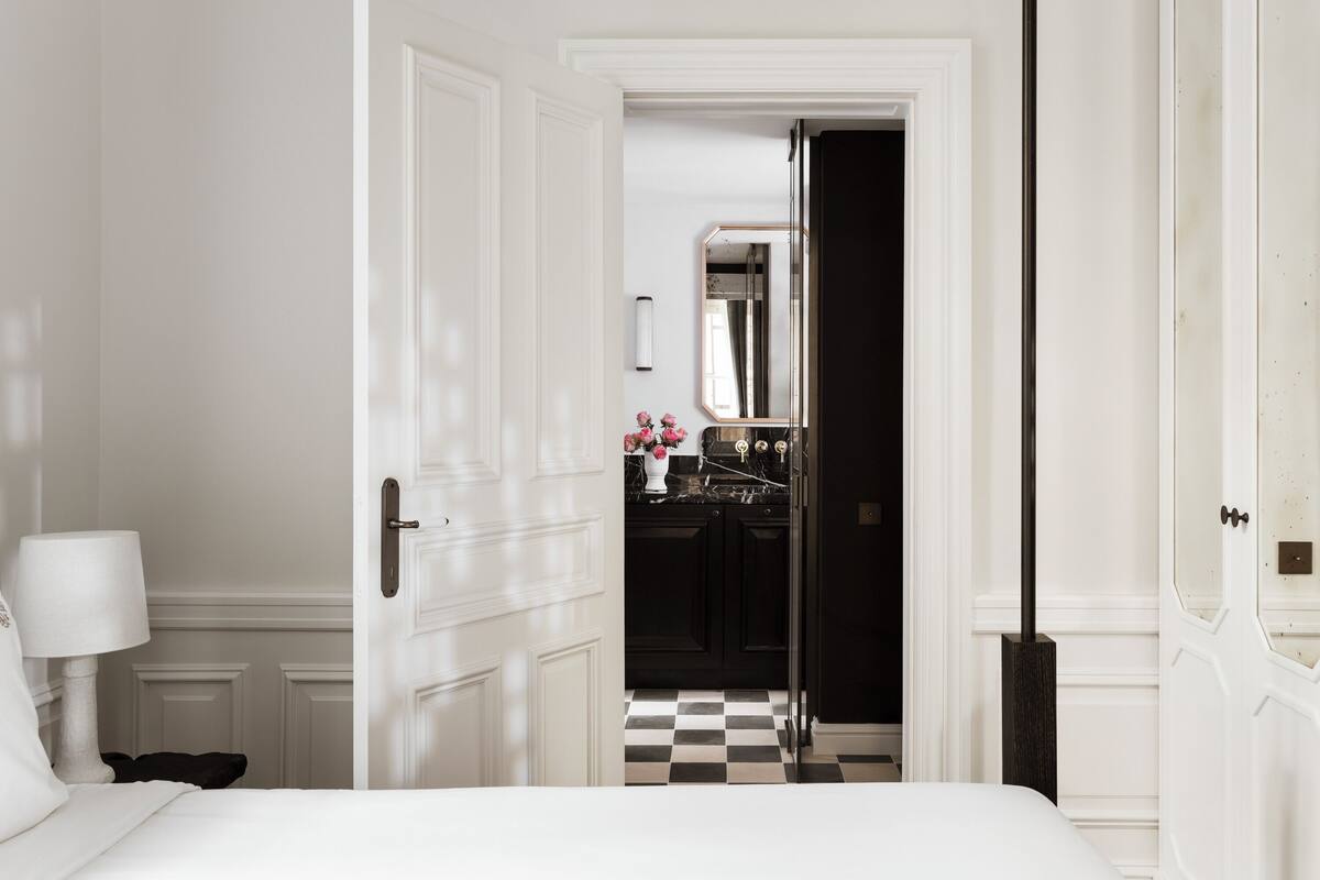 Quarto de hotel com piso xadrez, cama, mesa de cabeceira, abajur e porta que dá para um banheiro.