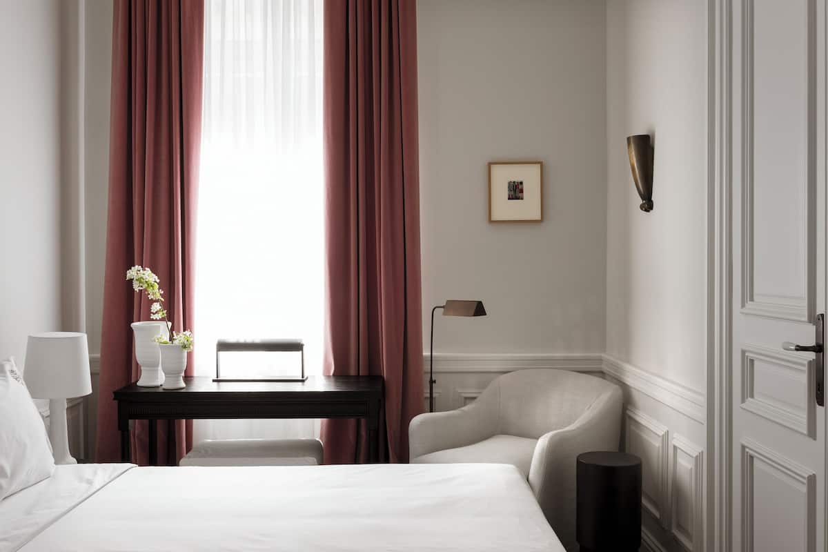 Quarto de hotel com cama, escrivaninha, cadeira, luminária, um quadro na parede e janela com cortinas.