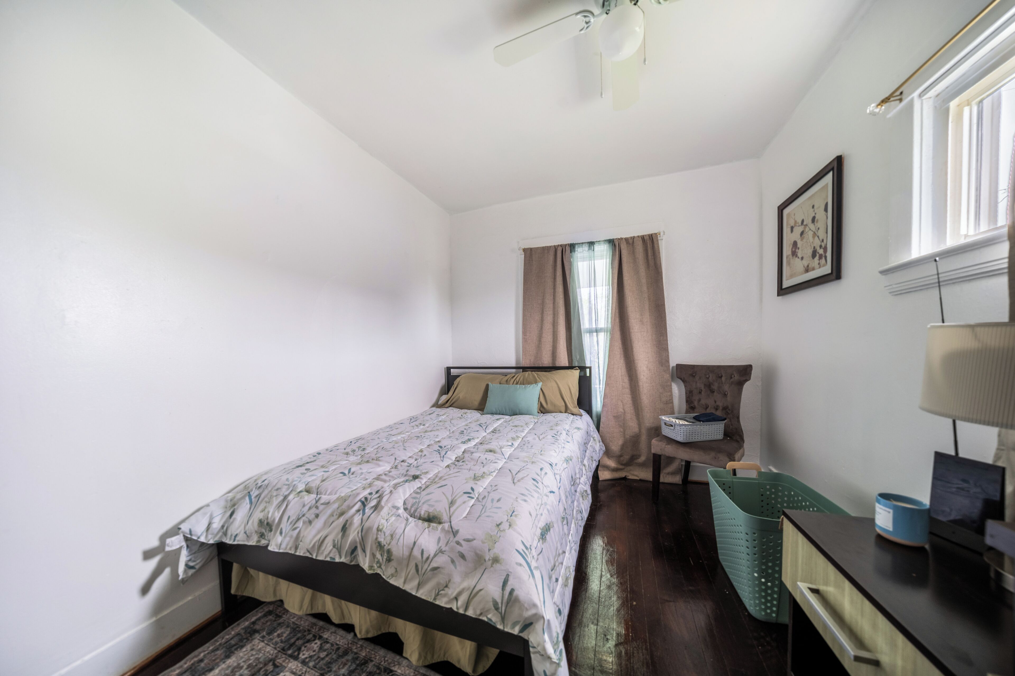 A bedroom with a bed, a chair, a desk, a lamp, a window with curtains, and a ceiling fan.
