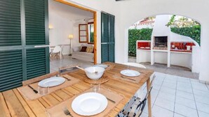 Ein Essbereich im Freien mit einem Holztisch für vier Personen, einem Grill und Blick auf einen Garten.