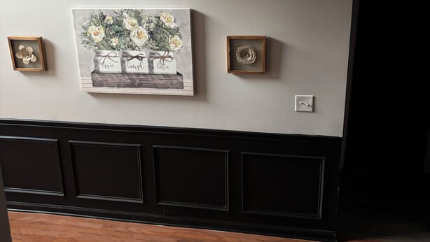 Eine Wand mit einem gerahmten Blumenbild, zwei kleinen gerahmten Objekten und einem Bereich mit schwarzem Wandpaneel.