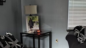 Ein Wohnzimmer mit grauem Sofa, einem kleinen Tisch mit Lampe, einem Bilderrahmen und einem Fenster mit Jalousien.