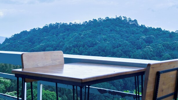 Een houten tafel en stoel op een balkon met uitzicht op een dichtbegroeide heuvel.