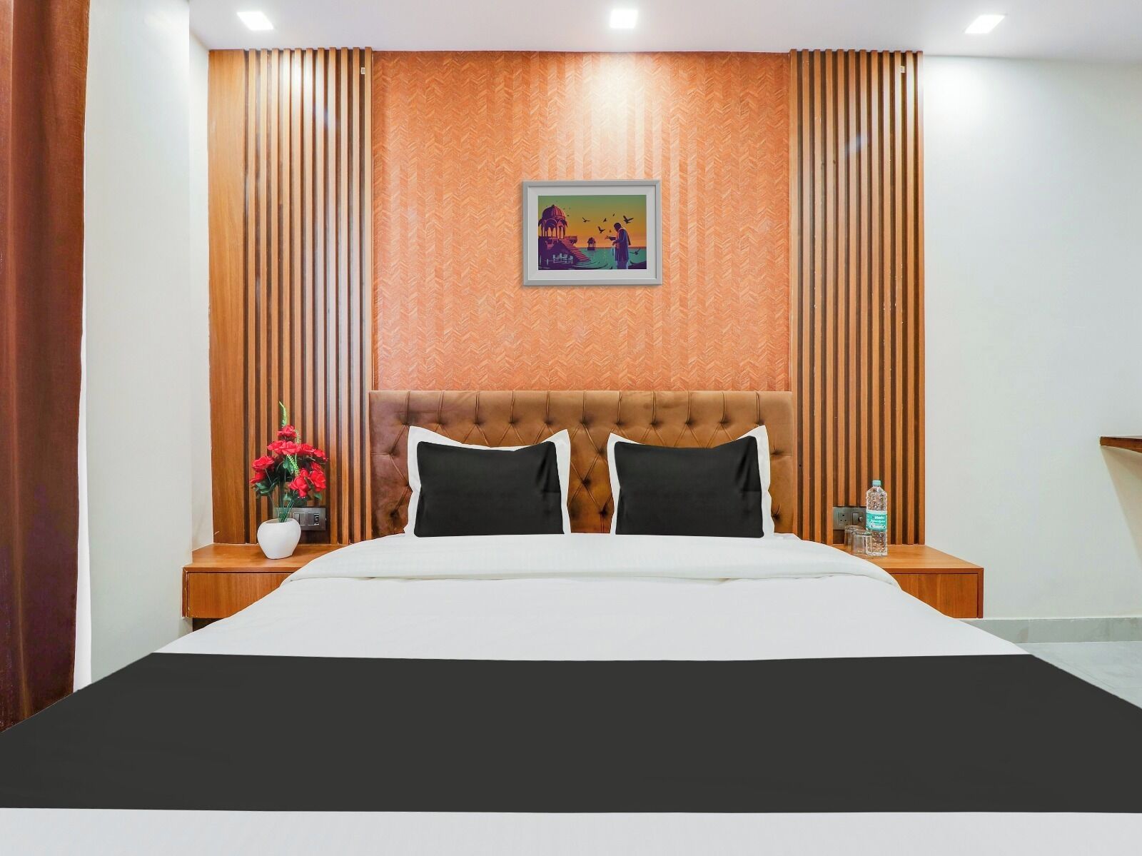 A hotel room with a large bed, wooden paneling on the walls, a framed picture on the wall, and a nightstand with a vase of flowers.