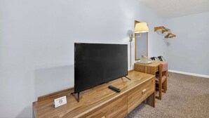 Une pièce comprenant un meuble TV en bois, un téléviseur à écran plat, une télécommande, une lampe et une petite table avec une chaise.