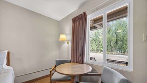 A hotel room with a round table, two chairs, a lamp, a window with curtains, and a view of trees.