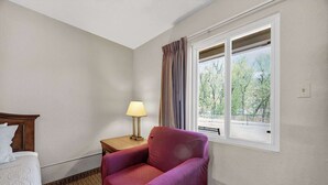 A hotel room with a bed, a chair, a nightstand with a lamp, a window with curtains, and a view of trees outside. - Eagle Motel Suite 118 (Manitou Springs)