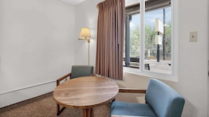 A room with a round wooden table, two chairs, a lamp, and a window with curtains. - Eagle Motel Suite 108 (Manitou Springs)