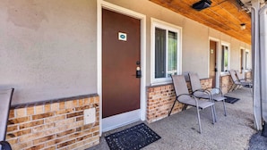 A building with a brown door, brick lower half, and a covered porch with chairs. - Eagle Motel Suite 108 (Manitou Springs)