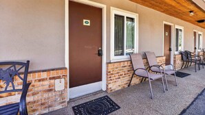 A building with a brown door, a brick wall, and a covered porch with chairs. - Eagle Motel - Suite 116 (Manitou Springs)