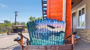 A wooden chair with a painted 'Eagle Motel' sign, a building with a brick facade, and a small structure in the background. - Eagle Motel - Suite 116 (Manitou Springs)