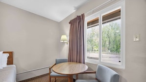 Quarto de hotel com uma cama, uma mesa redonda de madeira com duas cadeiras, uma luminária e uma janela com cortinas.