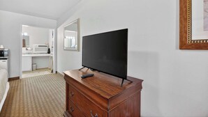 A hotel room with a wooden dresser, a television, a mirror, and a kitchenette in the background. - Eagle Motel Suite 117 (Manitou Springs)