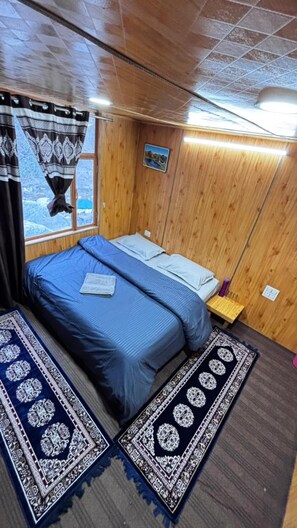 A wooden room with a bed, a window, a wall-mounted picture, and a small table. - Zenden Stay And Cafe (Jibhi)