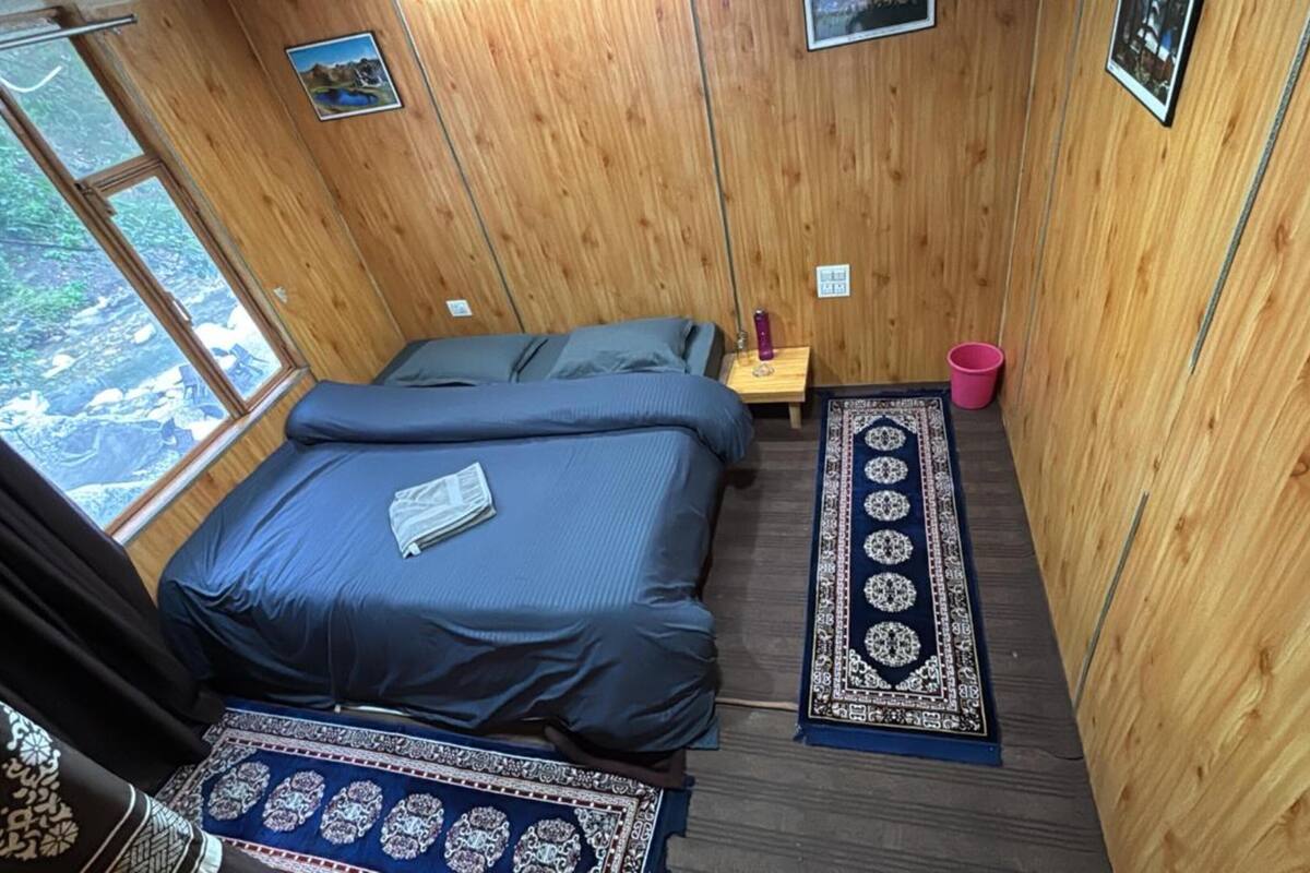 A wooden room with a bed, a nightstand, a small table, a rug, and a window with a view of greenery.