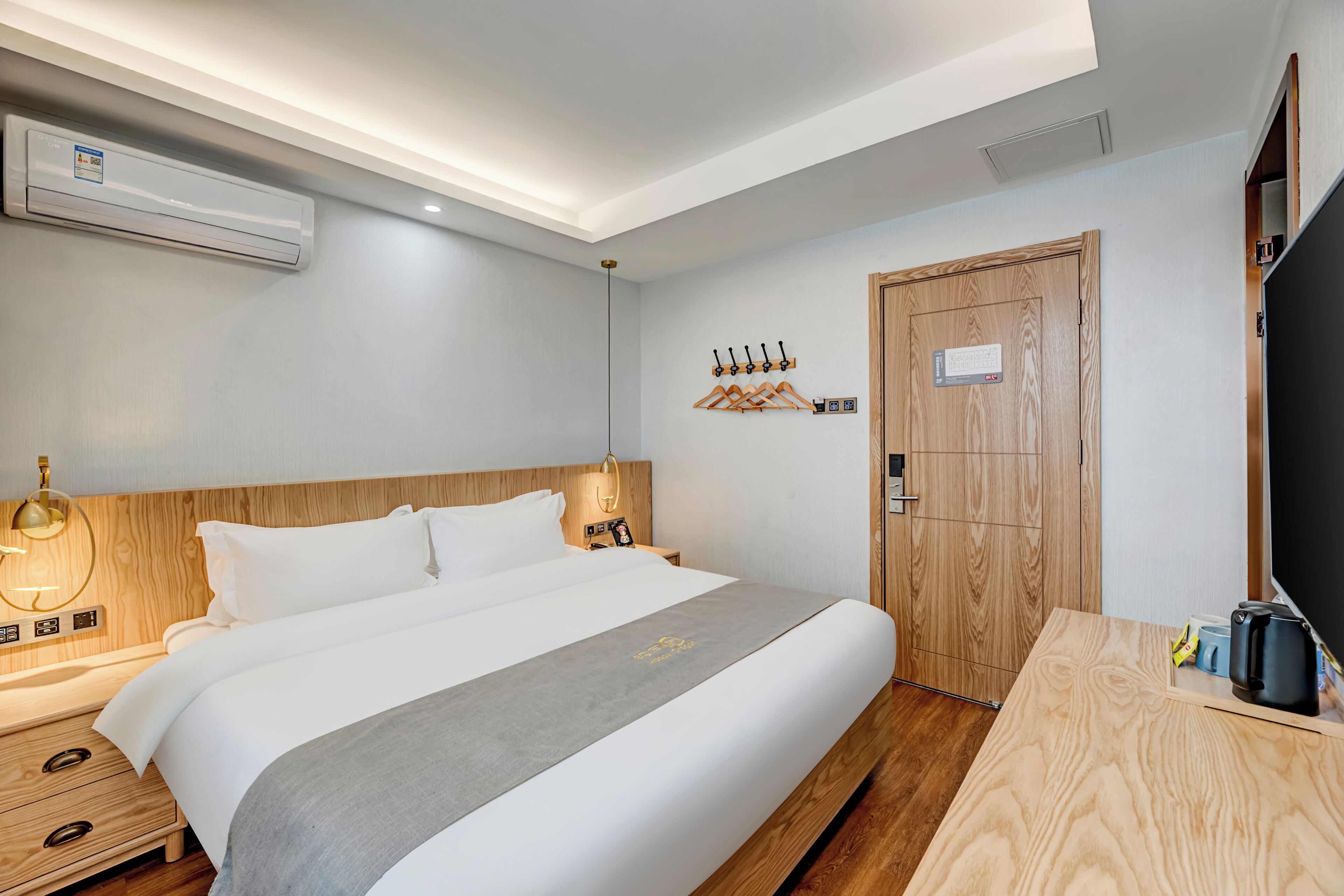 Une chambre d’hôtel comprenant un lit, une table de chevet, une lampe, un climatiseur mural, des patères et une porte en bois.