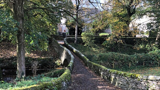 A stone pathway in a garden with a stone wall on one side and trees on the other. - Cross Winds (Lancaster)