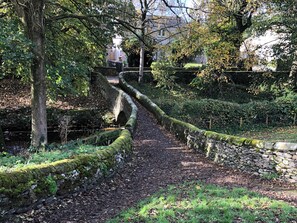 A stone pathway in a garden with a stone wall on one side and trees on the other. - Cross Winds (Lancaster)