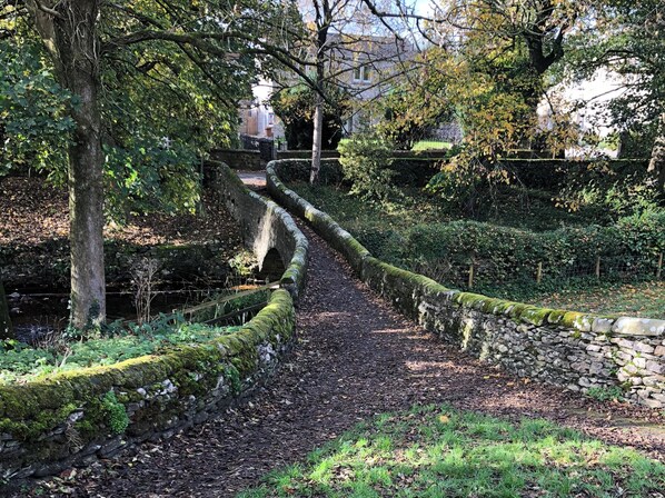 A stone pathway in a garden with a stone wall on one side and trees on the other. - Cross Winds (Lancaster)
