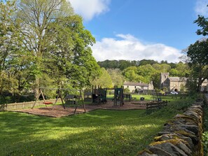 A park with playground equipment, a stone wall, and a building in the background. - Cross Winds (Lancaster)