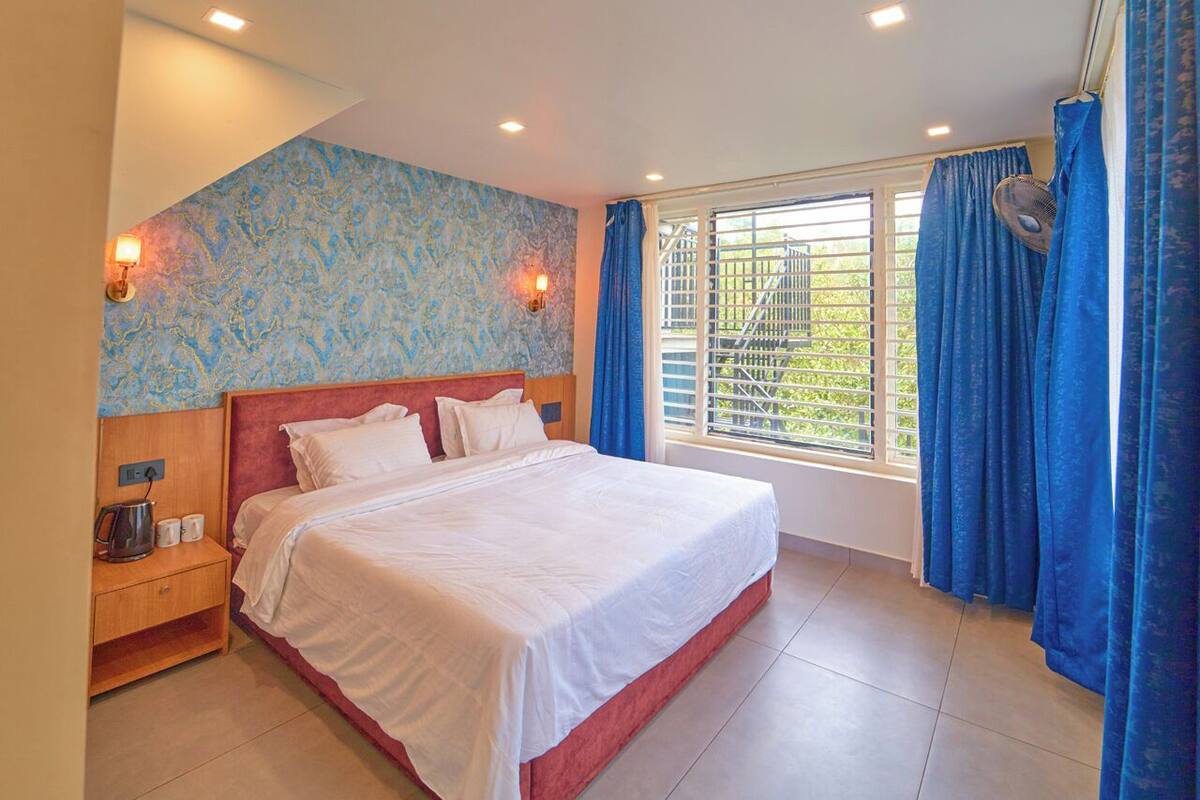 A bedroom with a large bed, a nightstand, a wall-mounted lamp, and a window with curtains.