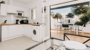 A modern kitchen with a washing machine, a microwave, and a coffee maker. There is a dining area with a table and chairs, and a view of a pool and greenery outside. - Spectacular apartment (Cap d'Artrutx)