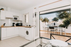 Een moderne keuken met een wasmachine, een magnetron en een koffiezetapparaat. Er is een eethoek met een tafel en stoelen, en uitzicht op een zwembad en groen buiten.