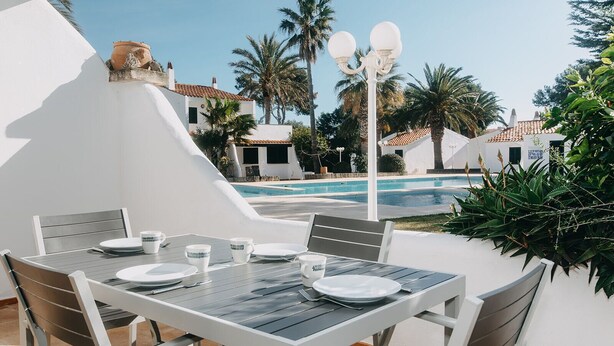 An outdoor dining area with a table set for four, surrounded by white walls and palm trees, overlooking a swimming pool. - Spectacular apartment (Cap d'Artrutx)