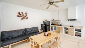Un espacio habitable funcional con sofá, mesa de comedor, sillas, ventilador de techo, zona de cocina y estante en la pared.