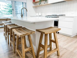 A modern kitchen with a white island, wooden stools, and a view of trees through the window. - Morgies on Long Beach (Long Beach)