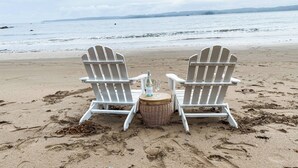Zwei weiße Adirondack-Stühle an einem sandigen Strand, mit einer Flasche und Gläsern auf einem kleinen runden Tisch.