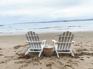 Zwei weiße Adirondack-Stühle an einem sandigen Strand, mit einer Flasche und Gläsern auf einem kleinen runden Tisch.