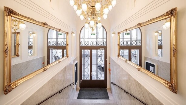 Een statige hal met sierlijke spiegelramen met gouden lijst, een kroonluchter en een dubbele deur met een glazen balustrade.