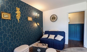 Una habitación con pared azul oscuro con estampado, un sofá azul con cojines, un reloj de pared redondo y una mesita con una maleta y un control remoto.