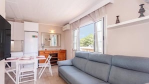 Ein Wohnzimmer mit grauem Sofa, einem Esstisch mit Stühlen, einer Küchenzeile inklusive Kühlschrank und einem Fenster mit Blick auf Bäume.