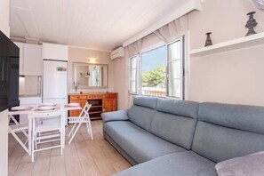 A living room with a grey sofa, a dining table with chairs, a kitchen area with a refrigerator, and a window with a view of trees. - Beautiful apartment Bon Repos 8 (Cap d'Artrutx)
