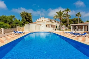 Una piscina con sillones de descanso, un edificio de techo rojo y palmeras al fondo.