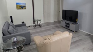 A living room with a grey sofa, a beige armchair, a glass coffee table, a TV on a stand, and a painting on the wall. - Spacious 3 Bedroom Private Guest Suite (Inuvik)