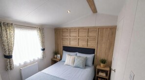 Ein Schlafzimmer mit Holz-Kopfteil, einem Bett mit vielen Kissen, einem Nachttisch mit Pflanze und einem Fenster mit Vorhängen.