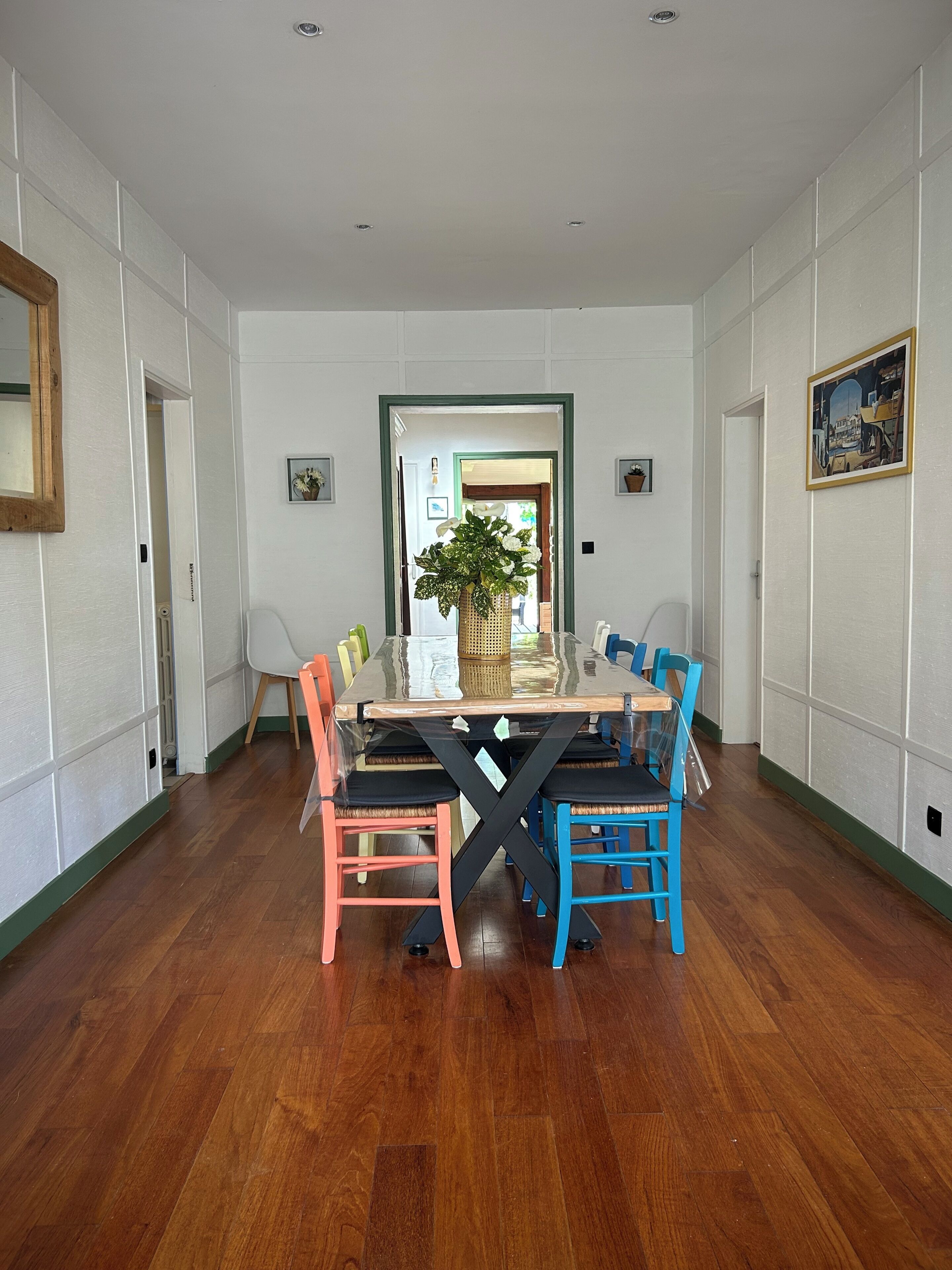 Un comedor con una mesa de madera y sillas coloridas, una planta en maceta y un cuadro en la pared.