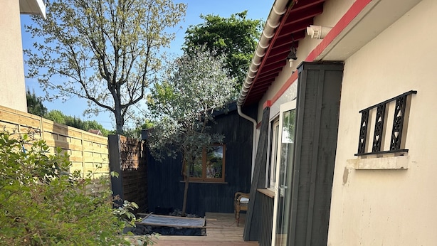 Een houten terras leidt naar een huis met een raam en een deur, omringd door groen.
