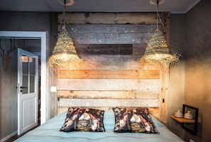 Un dormitorio con cabecera de madera, dos lámparas colgantes, una cama con almohadas estampadas y un pequeño estante con adornos.