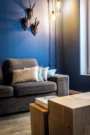 A modern living room with a dark blue wall, a gray sofa with two pillows, a wooden coffee table, and three mounted animal skulls with hanging light bulbs. - House - Urban decor gîte, La Luciole, ideally located in the heart of the Ardennes, (Marche-en-Famenne)